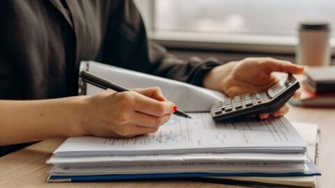A woman's hand holding a pen and calculator on top of a stack of paperwork