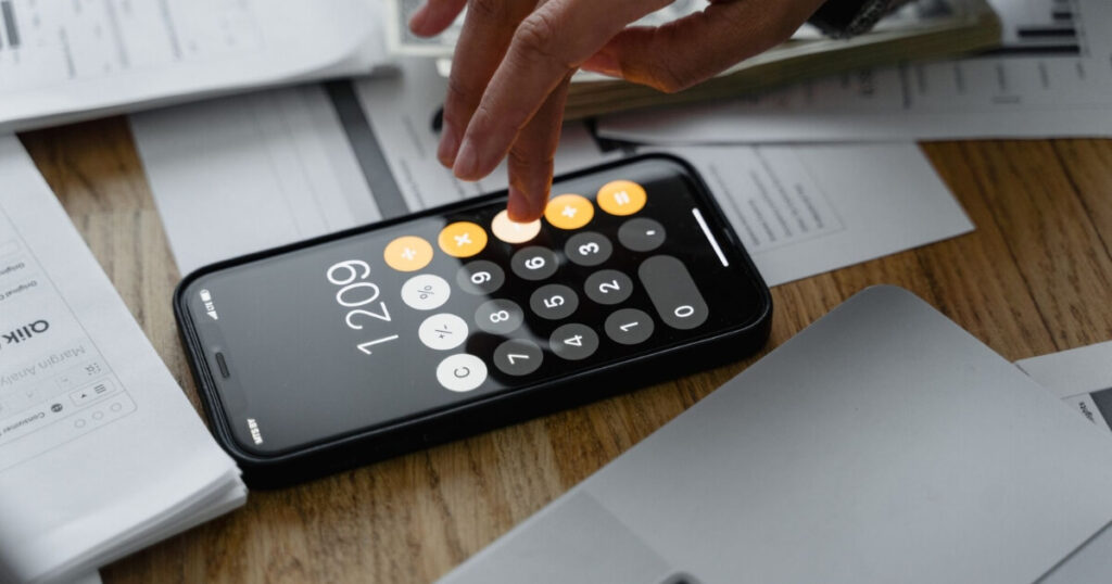 A finger tapping a button on the calculator app on the iPhone, surrounded by paperwork on a table