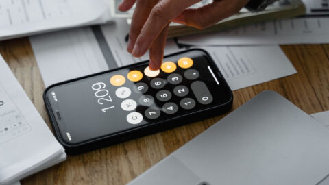 A finger tapping a button on the calculator app on the iPhone, surrounded by paperwork on a table