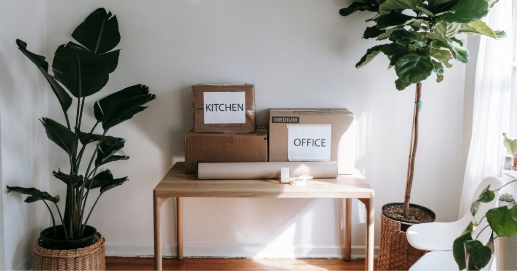 Stylish interior with moving boxes labeled 'Kitchen' and 'Office' on a wooden table surrounded by houseplants