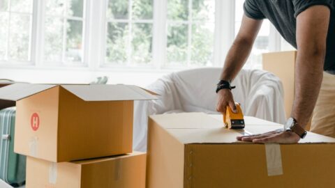 Man sealing cardboard boxes in a bright modern living room, preparing for a move