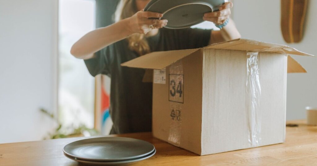 A woman unpacks ceramic plates from a cardboard box on a wooden table in a cozy indoor setting