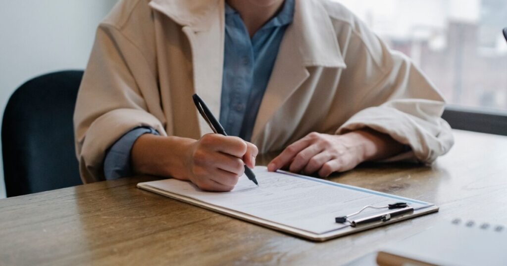 A woman in a formal setting fills out paperwork on a clipboard at an office desk
