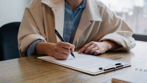 A woman in a formal setting fills out paperwork on a clipboard at an office desk