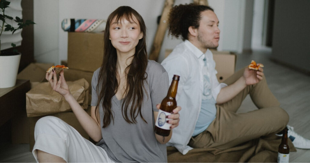 Two renters happily enjoy pizza and beer while relaxing indoors with moving boxes