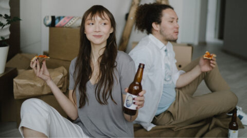 Two renters happily enjoy pizza and beer while relaxing indoors with moving boxes