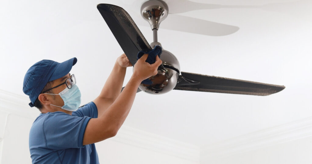 A man wearing a face mask and cleaning a ceiling fan with a cloth
