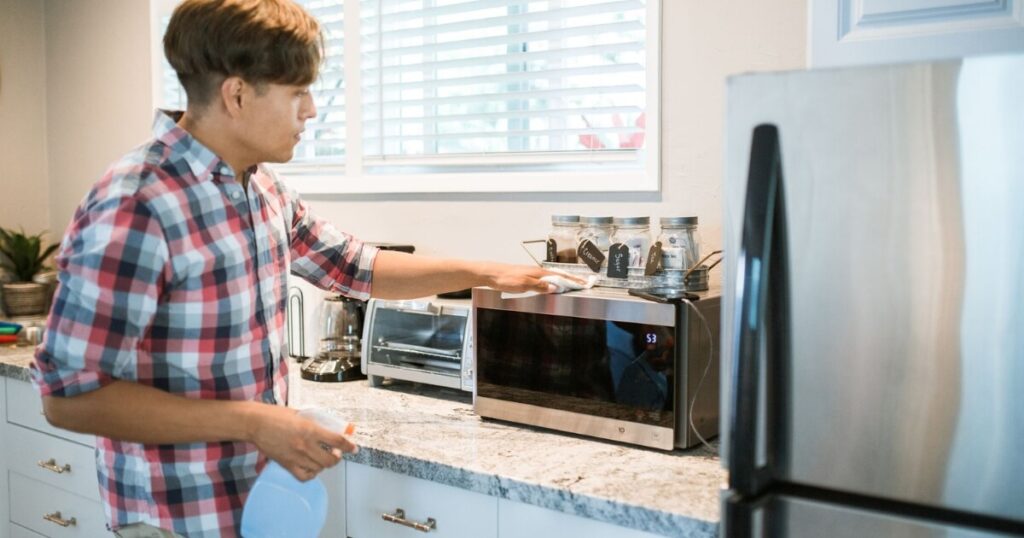 A man is cleaning a microwave in his modern kitchen using a spray bottle and cloth