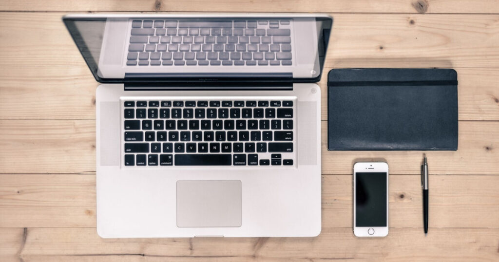 Top view of a tidy workspace featuring a laptop, smartphone, notebook, and pen on a wooden desk