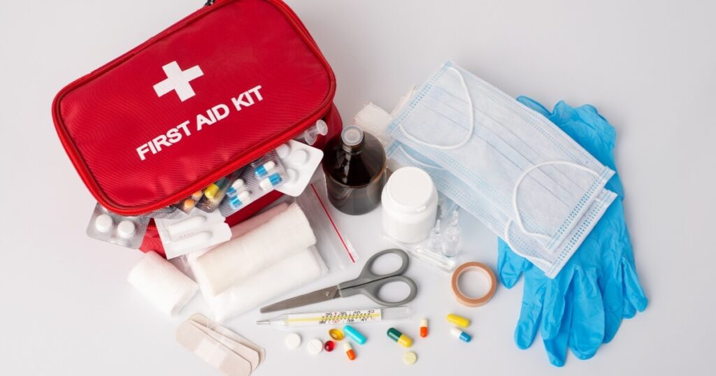 Open red first aid pouch surrounded by bandages, medical scissors, medication, face masks, and blue disposable gloves on a white background.