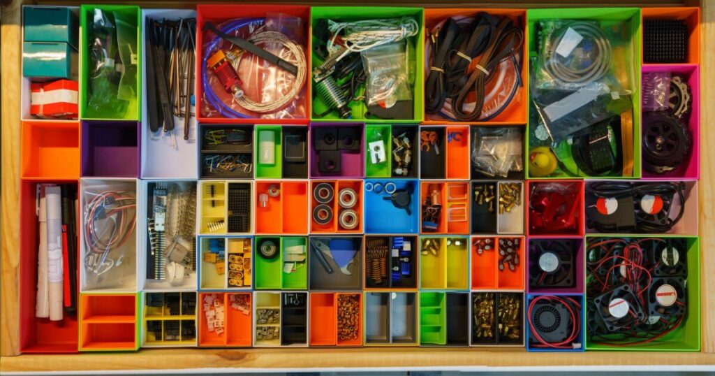Top-down view of a neatly organized drawer filled with various small items, hardware, tools, and cables sorted into small, colorful rectangular compartments.
