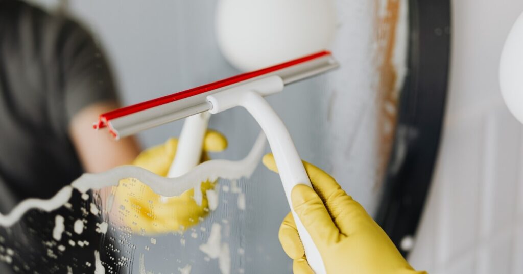 Person wearing yellow rubber gloves using a white and red squeegee to wipe soapy foam off a bathroom mirror.